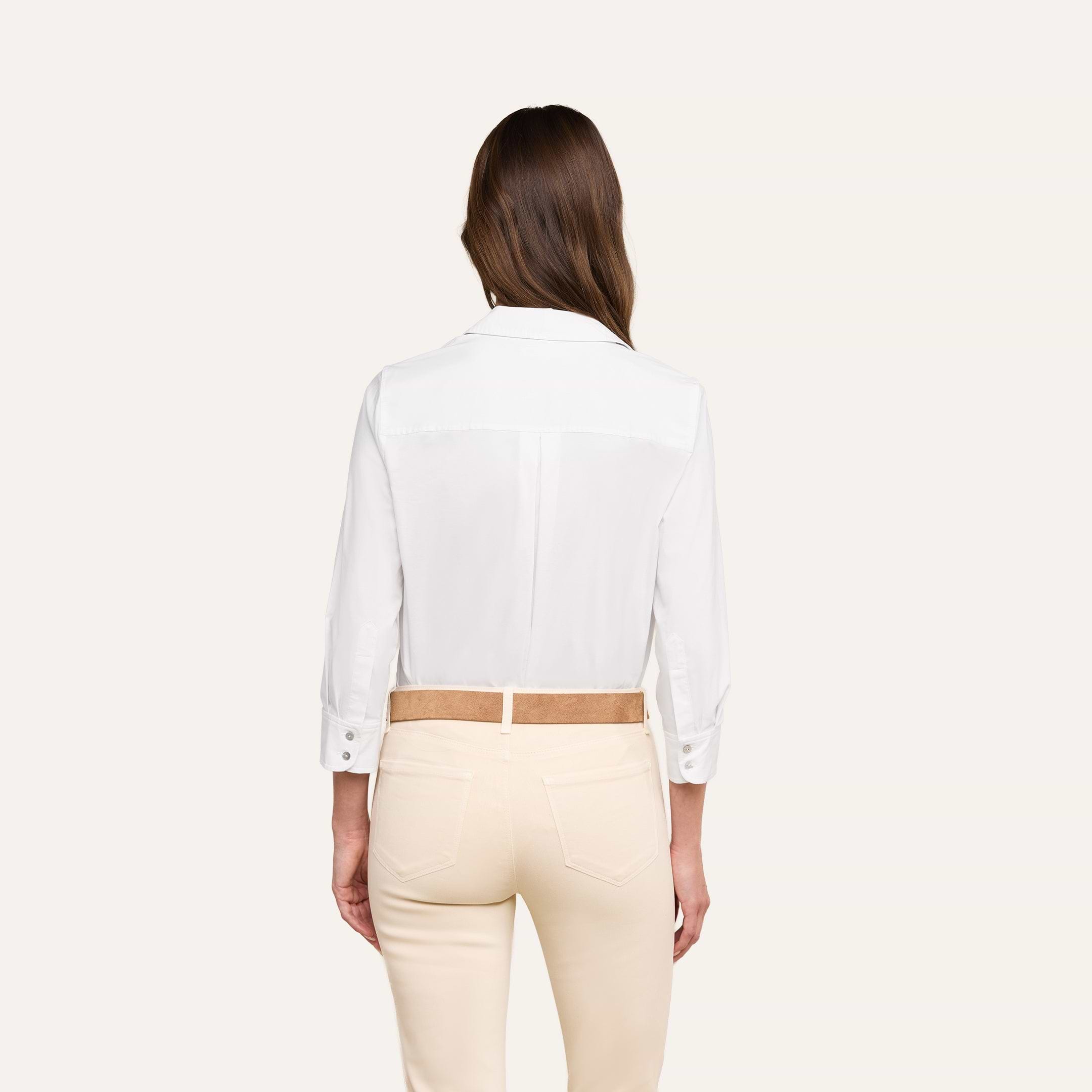 Woman standing with her back to the camera, wearing the Daniella Cotton-Blend Blouse—a relaxed fit white stretch cotton poplin long-sleeve shirt—tucked into high-waisted, light beige pants with a tan belt, against a plain off-white background.