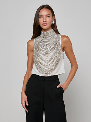 A woman wears the Yun Draped Crystal Tank with layered silver chains and black high-waisted pants, standing against a light gray background, one hand in her pocket—an effortlessly chic holiday party look.