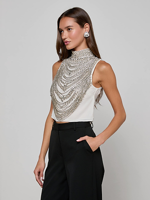 A woman with long brown hair wears the Yun Draped Crystal Tank featuring layered silver chains, paired with black high-waisted pants and large silver earrings. She stands against a light gray background—an ideal outfit for holiday parties.