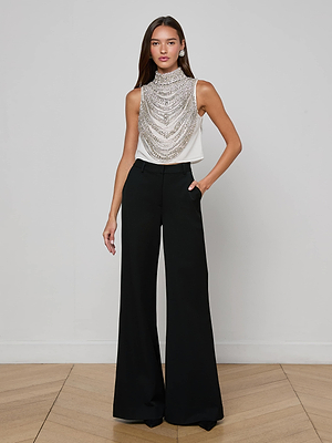 A woman with long straight hair stands against a plain background wearing the Yun Draped Crystal Tank—sleeveless, embellished with layers of silver chains—paired with high-waisted, wide-leg black trousers for a chic holiday look. One hand rests in her pocket.