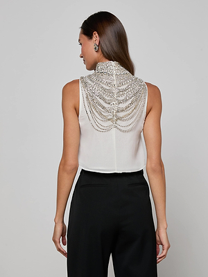 A woman facing away wears the Yun Draped Crystal Tank, featuring intricate silver beadwork and crystals on the back, paired with high-waisted black pants. Her straight brown hair and stud earrings complete this chic holiday party look.