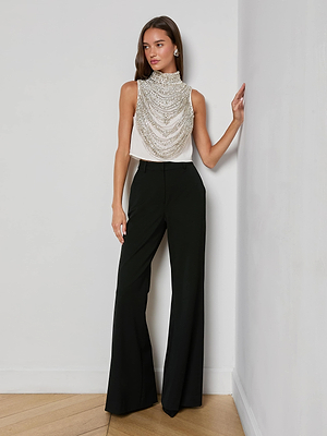 A woman with straight brown hair poses indoors by a white wall, wearing the Yun Draped Crystal Tank paired with high-waisted, wide-leg black trousers—a chic look perfect for holiday parties. One hand rests on the wall as she shows off her stylish outfit.
