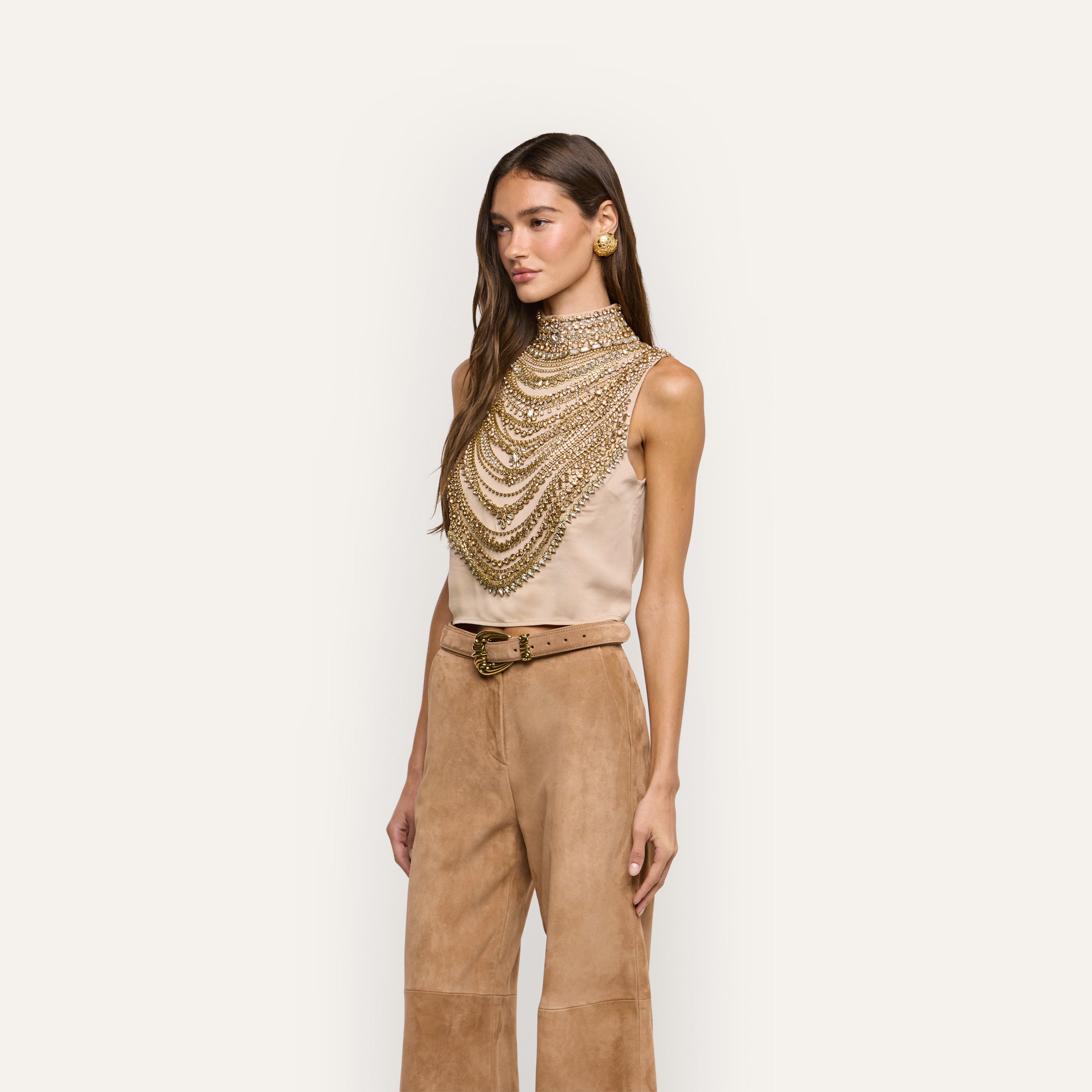 The Yun Crystal Tank is a sleeveless beige statement top featuring a high neck and multiple layers of draped gold chains.