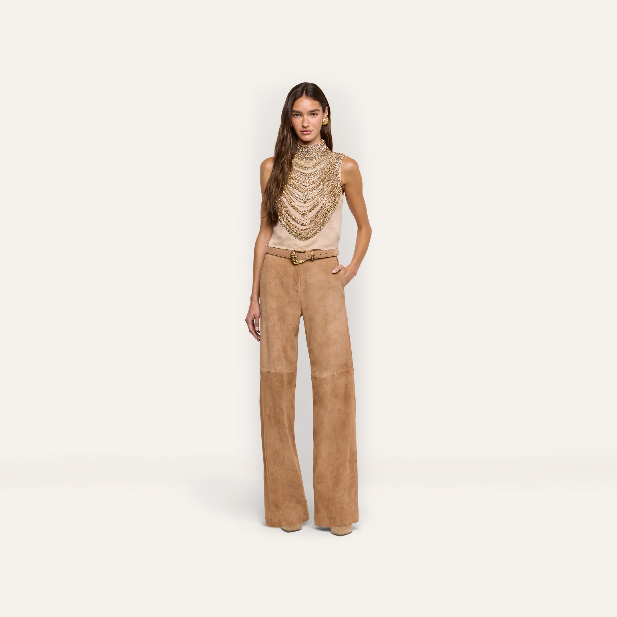 The Yun Crystal Tank is a sleeveless beige top featuring a high neckline with dangling gold crystals, worn with tan suede wide-leg trousers and a coordinating belt, perfect for holiday party wear.