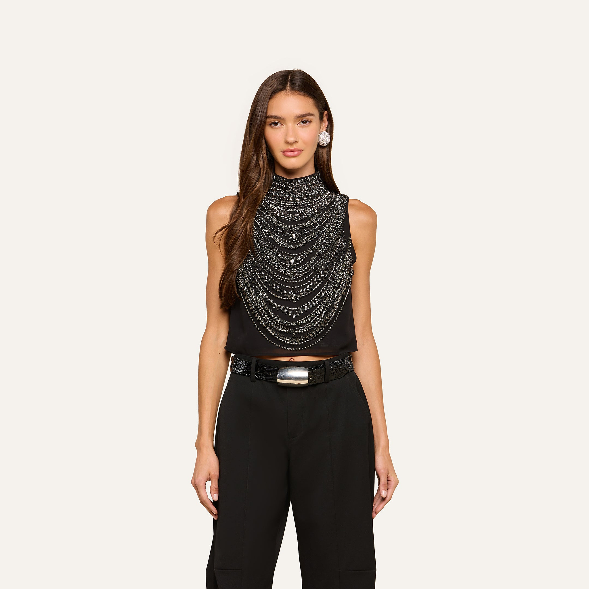 A woman with long brown hair wears the Yun Draped Crystal Tank, layered with silver chains, paired with black pants, a wide black belt with a silver buckle, and silver earrings, standing against an off-white background.