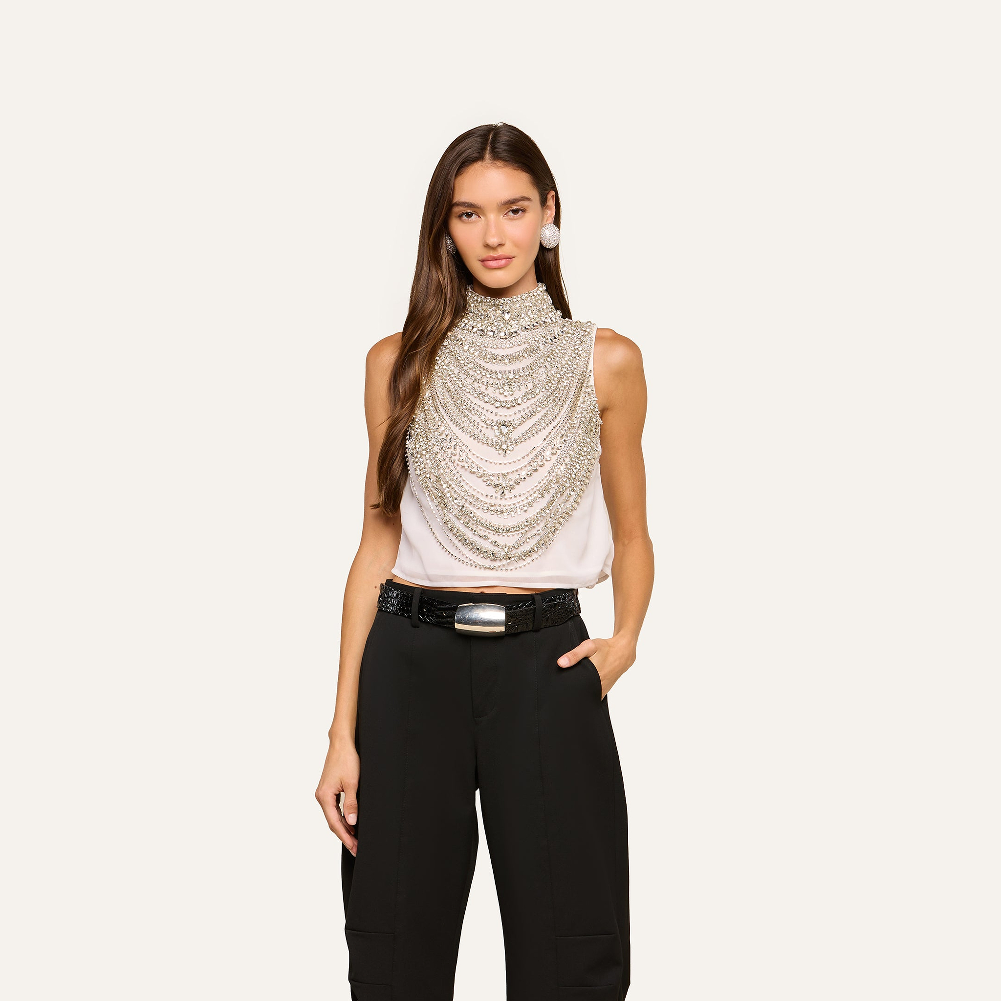 Wearing the Yun Draped Crystal Tank, layered silver chains, black high-waisted pants, a wide belt with a silver buckle, and large earrings, a woman stands against a plain light background.