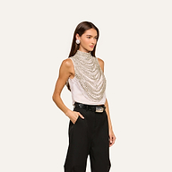 A woman stands with her hand in her pocket, wearing the Yun Draped Crystal Tank—a sleeveless statement ivory top adorned with layered silver chains—paired with black high-waisted pants, a black belt, and large round earrings against a plain light background. This holiday-ready piece elevates any outfit with its elegant sparkle.