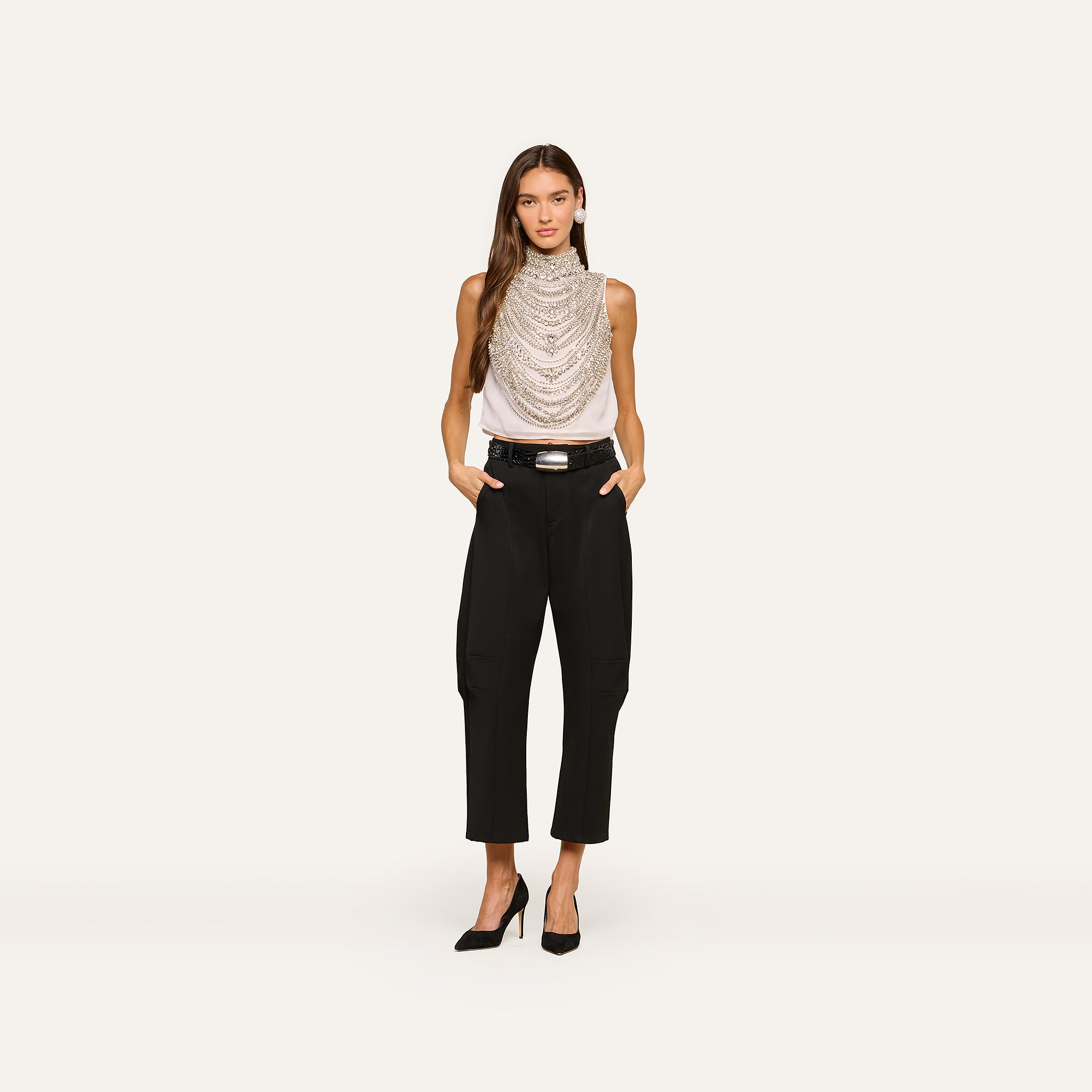 A woman stands against a plain background wearing the Yun Draped Crystal Tank styled with layered necklaces, black belted pants, and black heels, her hands in her pockets.