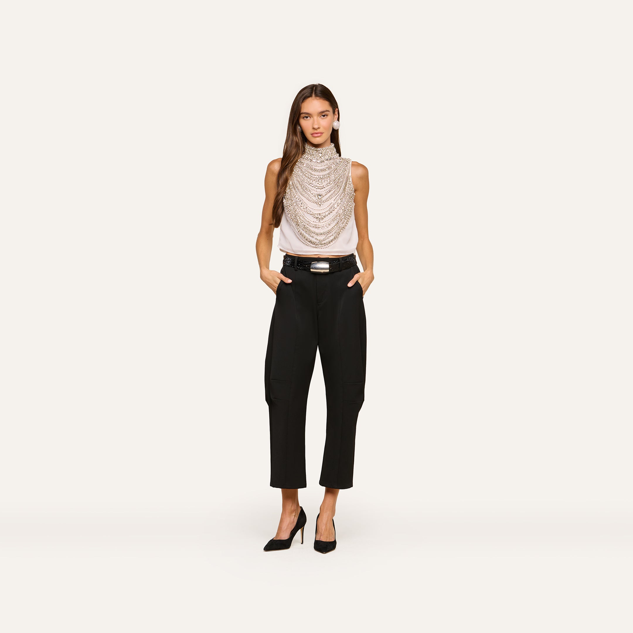 A woman stands against a plain background wearing the Yun Draped Crystal Tank styled with layered necklaces, black belted pants, and black heels, her hands in her pockets.