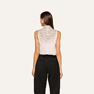 A woman stands with her back to the camera, wearing the Yun Draped Crystal Tank—a sleeveless ivory top featuring intricate beaded detailing on the back—paired with high-waisted black pants, set against a plain light background.