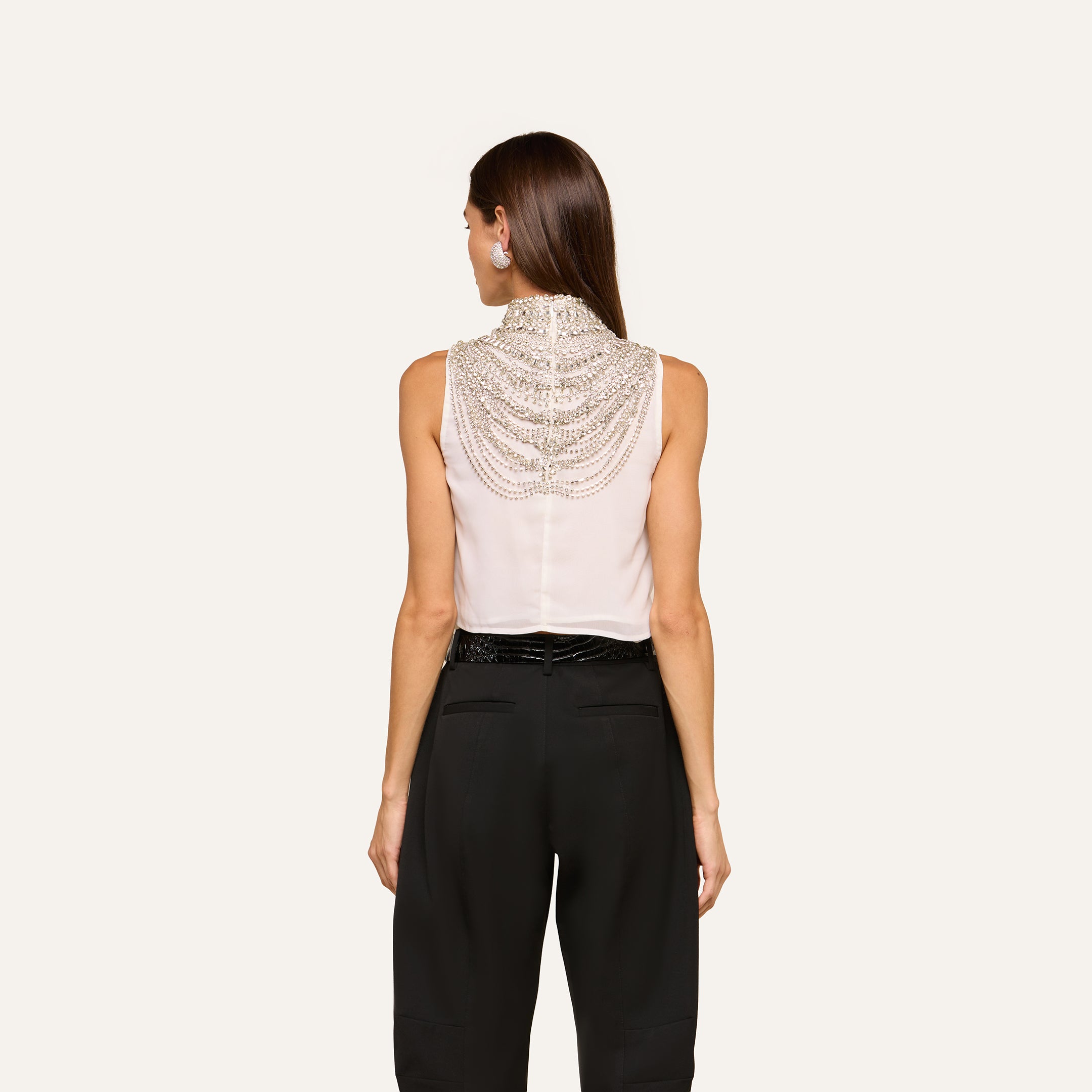 A woman stands with her back to the camera, wearing the Yun Draped Crystal Tank—a sleeveless ivory top featuring intricate beaded detailing on the back—paired with high-waisted black pants, set against a plain light background.