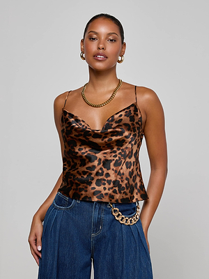 A woman wears the Calista Silk Camisole by L’AGENCE in leopard print, paired with a gold necklace, hoop earrings, wide-leg blue jeans, and a gold chain belt against a plain light gray background.