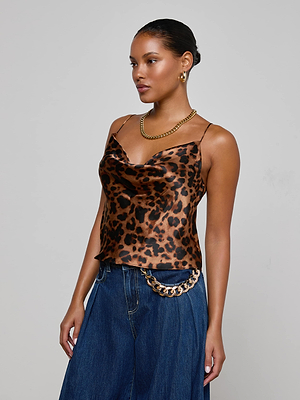 A woman stands against a light gray background wearing the L'AGENCE Calista Silk Camisole in leopard print, paired with wide-leg blue jeans, gold hoop earrings, a gold chain necklace, and a matching gold chain belt.