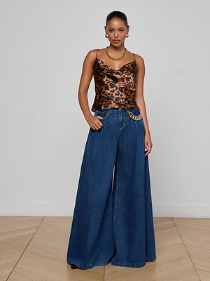 A woman stands against a plain wall wearing the L'AGENCE Calista Silk Camisole, wide-leg high-waisted blue jeans with a gold chain belt, gold jewelry, and hoop earrings. Her hair is pulled back with one hand in her pocket.