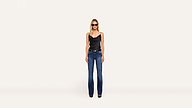 A woman wearing sunglasses, a black sleeveless top, blue jeans, and black shoes poses confidently against a minimalist white background.