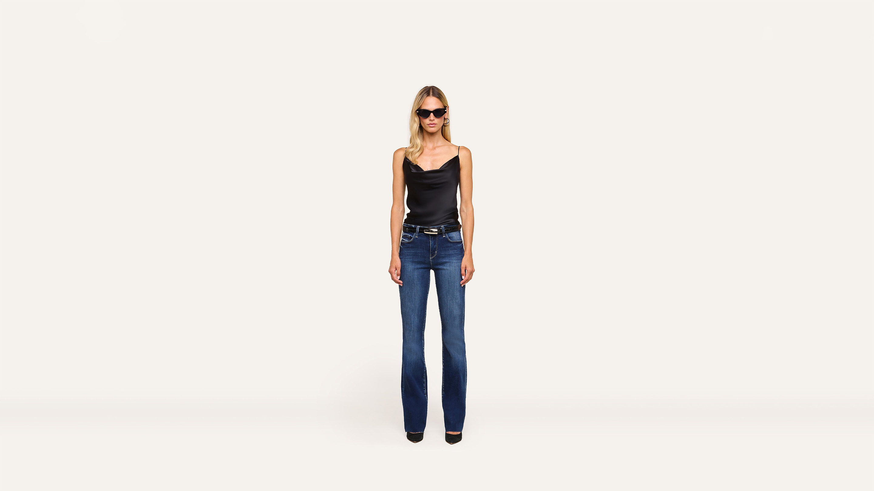 A woman wearing sunglasses, a black sleeveless top, blue jeans, and black shoes poses confidently against a minimalist white background.