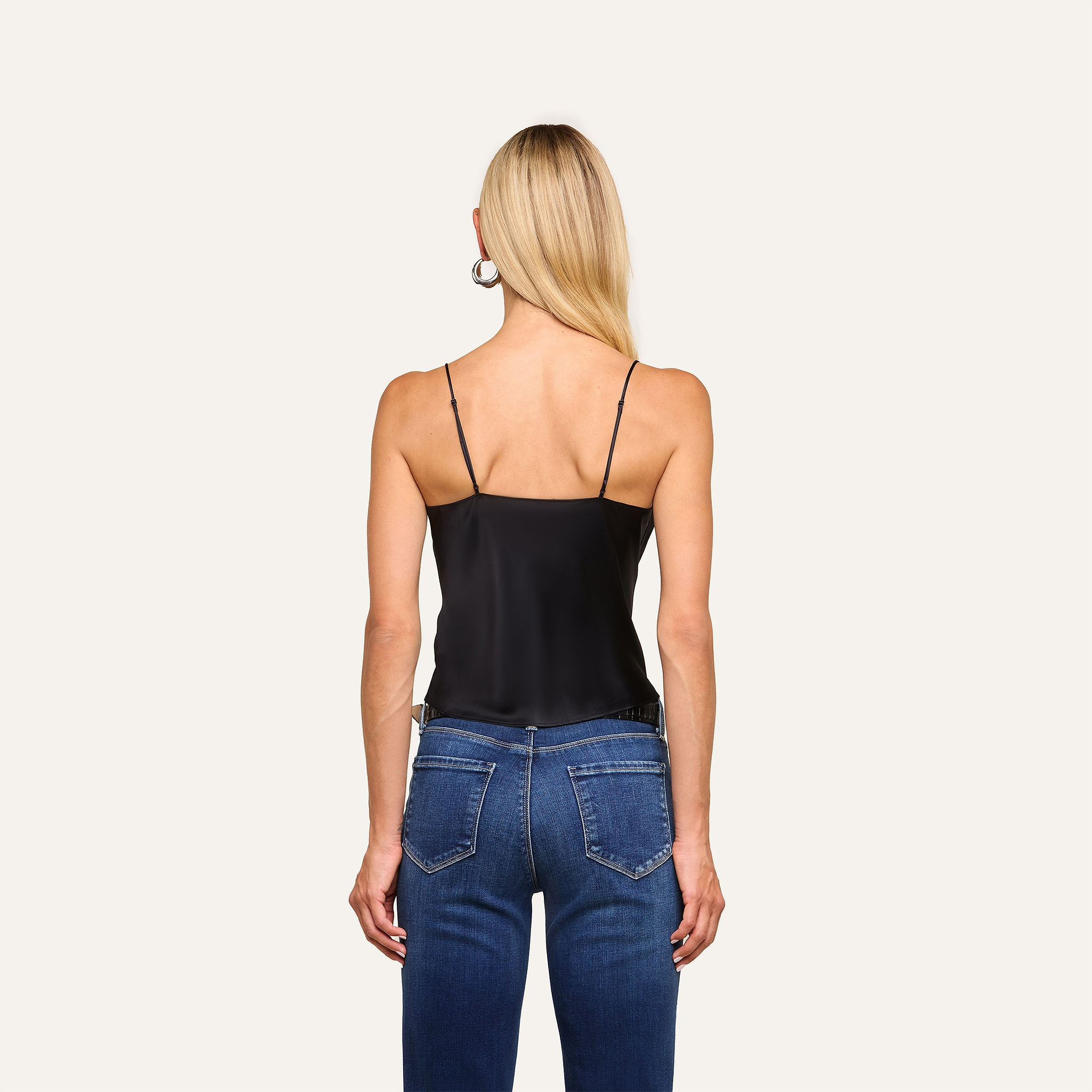 A woman with long blonde hair faces away, wearing the Calista Silk Camisole in black and blue jeans, standing against a plain light background.