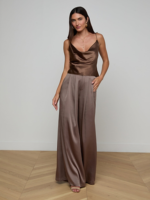 A woman stands indoors in the Calista Silk Camisole in brown with matching wide-leg pants. She has long dark hair, minimal jewelry, and neutral sandals, posing against a plain light wall and wooden floor.
