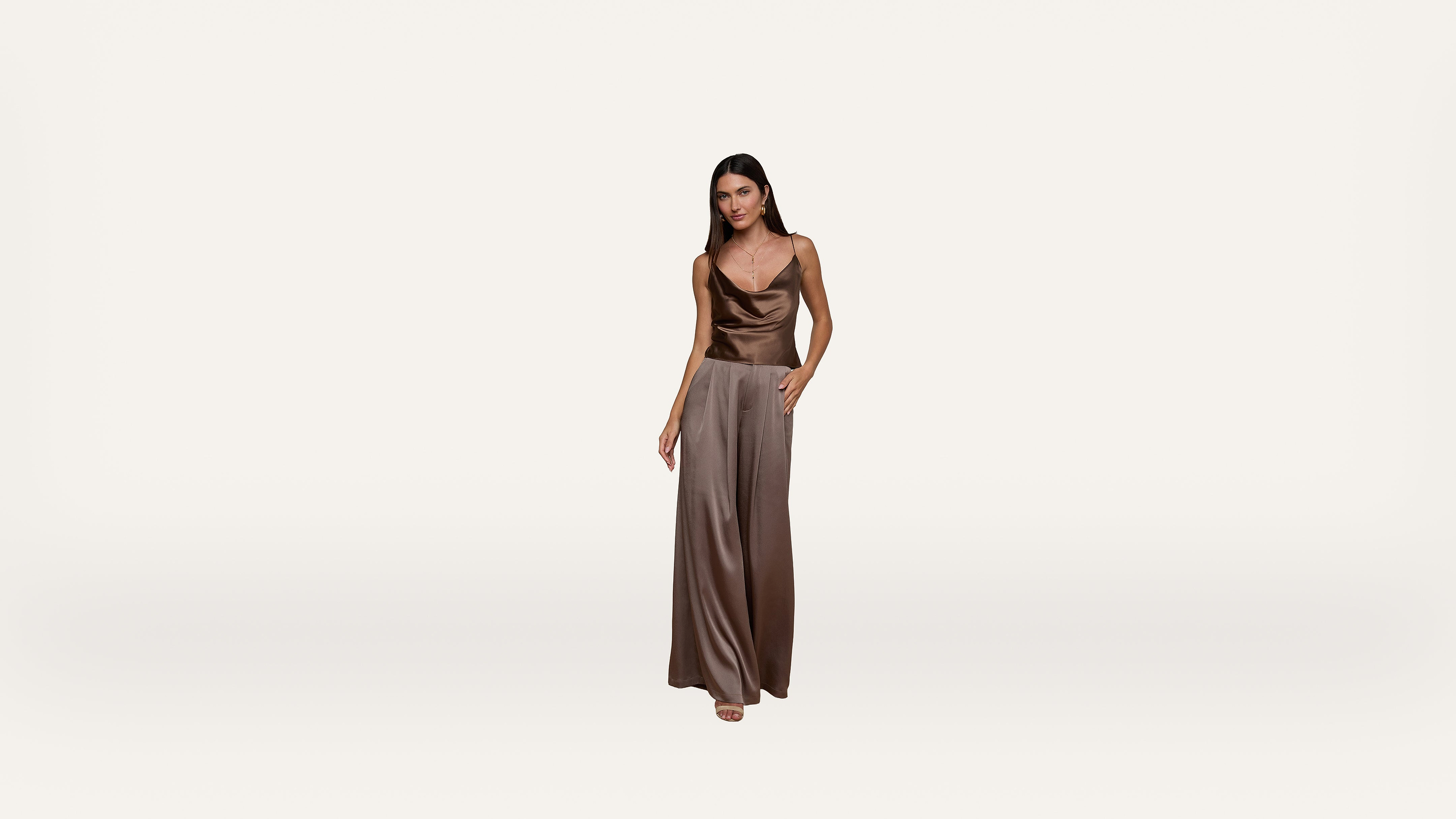Woman in a sleeveless, brown satin dress stands against a plain white background, facing forward with one hand in her pocket and the other relaxed by her side.