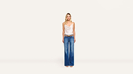 Woman standing against a plain white background, wearing a white sleeveless top, blue flared jeans, brown belt, and sandals. She has long blonde hair and a neutral expression, capturing a relaxed style.