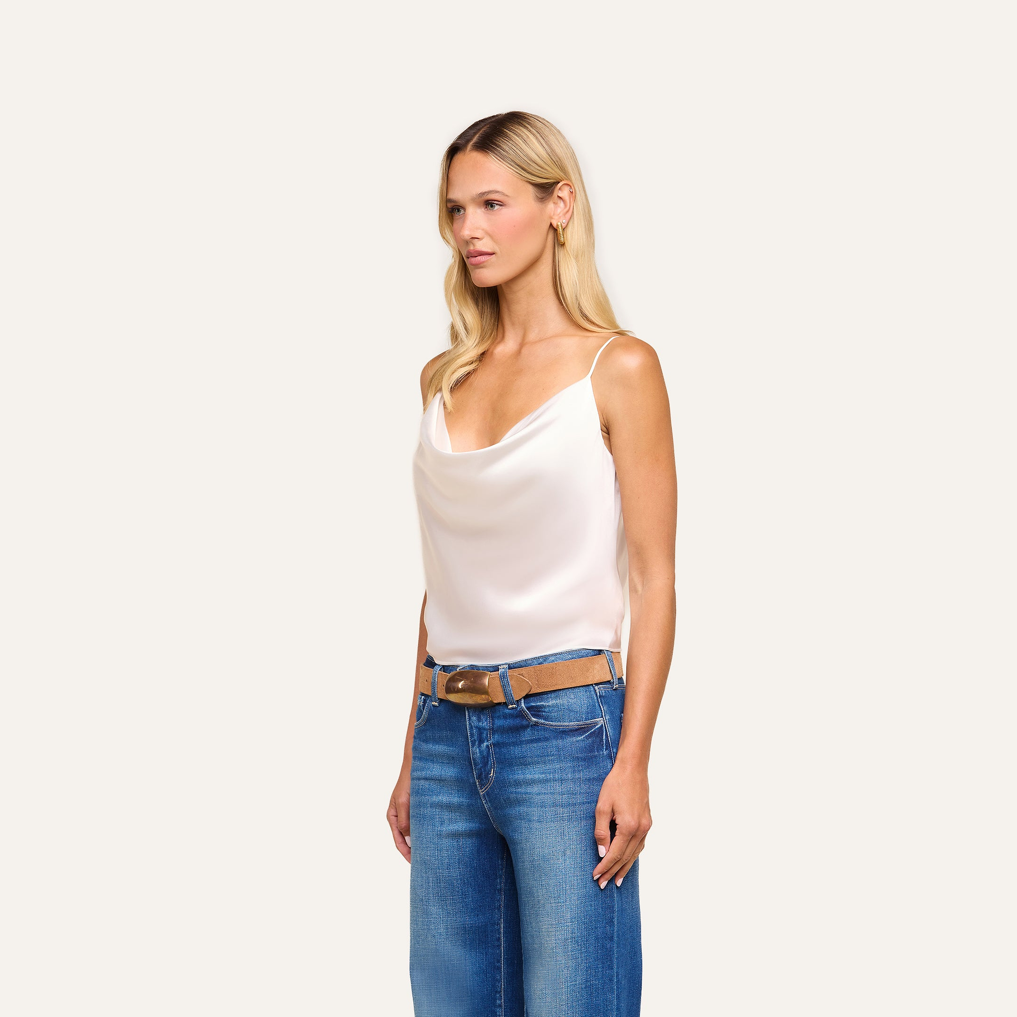 A woman with long blonde hair wears the Calista Silk Camisole in white, paired with blue jeans and a brown belt, standing against a plain light background.