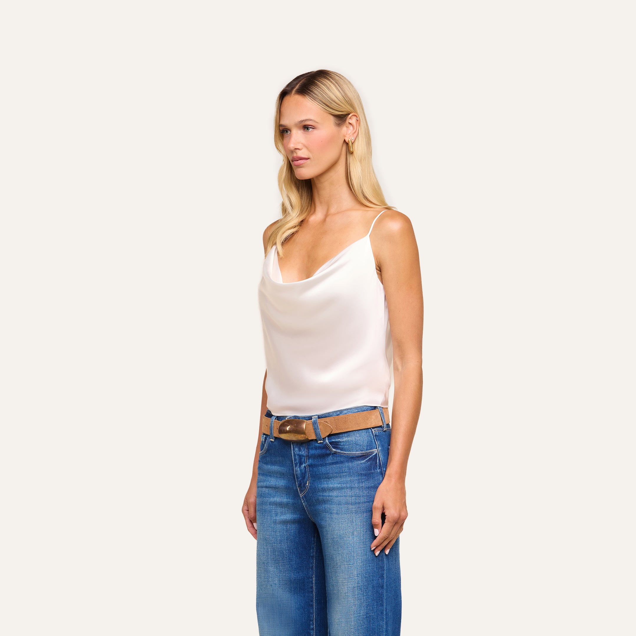A woman with long blonde hair wears the Calista Silk Camisole in white, paired with blue jeans and a brown belt, standing against a plain light background.