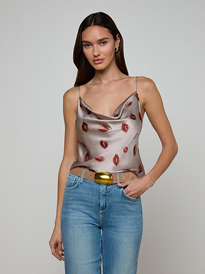 A woman with long brown hair wears the Calista Satin Camisole—silver with a red lip print and adjustable straps—paired with blue jeans and a gold belt, standing against a plain light gray background.