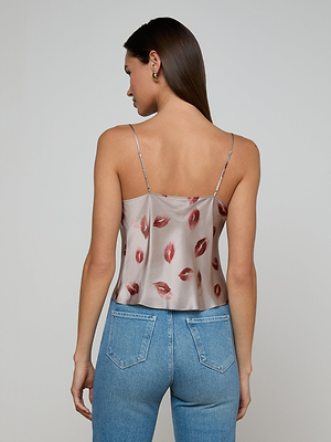 A woman with straight brown hair stands against a plain light gray background, wearing the Calista Satin Camisole in a cropped style with adjustable straps and red lip print, paired with light blue high-waisted jeans.