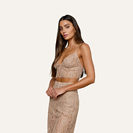 A woman with long brown hair wears the Aislin Lace Bralette in light beige, which features scalloped edges, paired with a matching lace skirt made from sheer stretch lace. She stands against a plain light background, looking at the camera with a neutral expression.