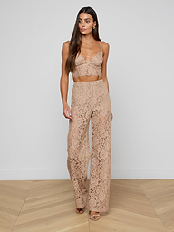 A woman wearing the Aislin Lace Bralette in beige with adjustable straps and matching high-waisted lace pants, both featuring sheer stretch lace and scalloped edges, stands against a plain white background.