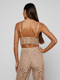 A woman with long brown hair stands with her back to the camera against a plain light background, wearing the Aislin Lace Bralette in beige—a sheer stretch lace bralette with adjustable straps—paired with matching lace pants.