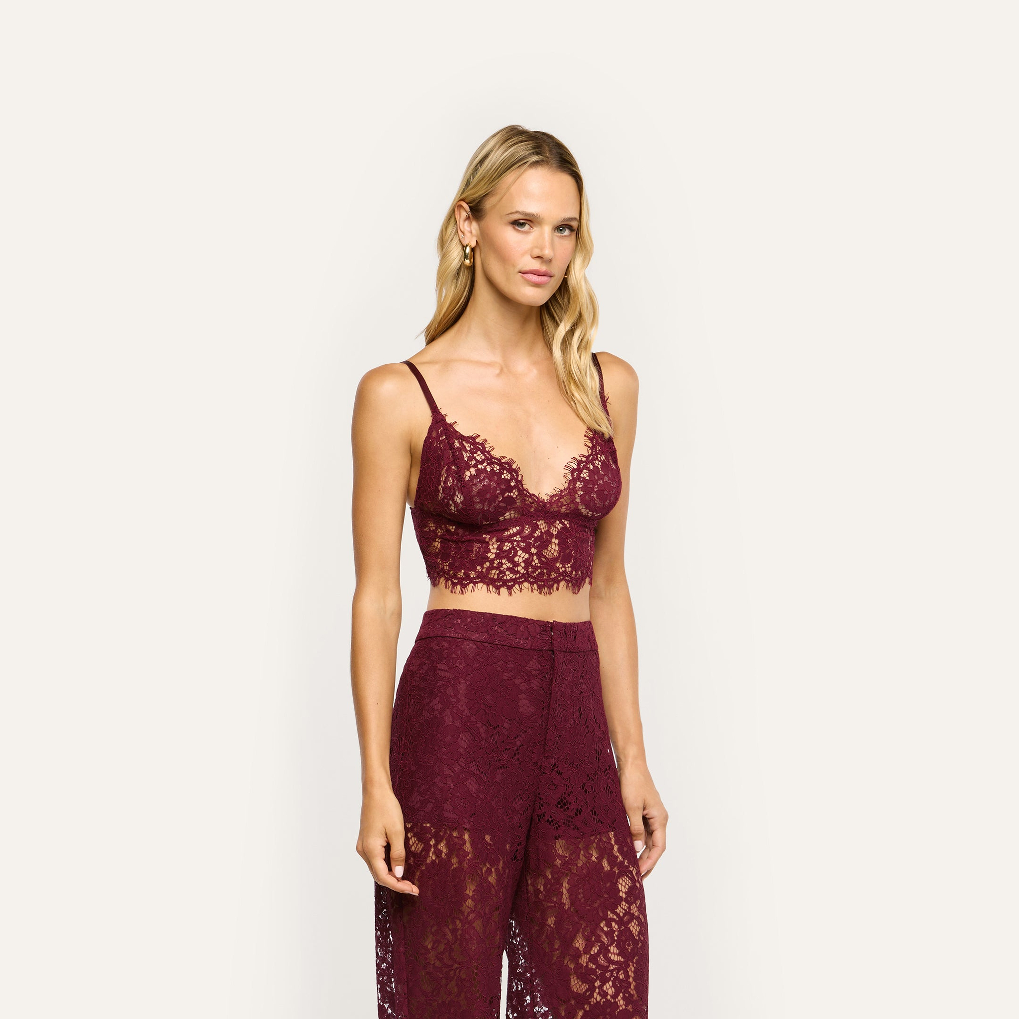 A woman wearing a deep red sheer stretch lace Aislin Lace Bralette with adjustable straps and matching high-waisted lace trousers with scalloped edges.