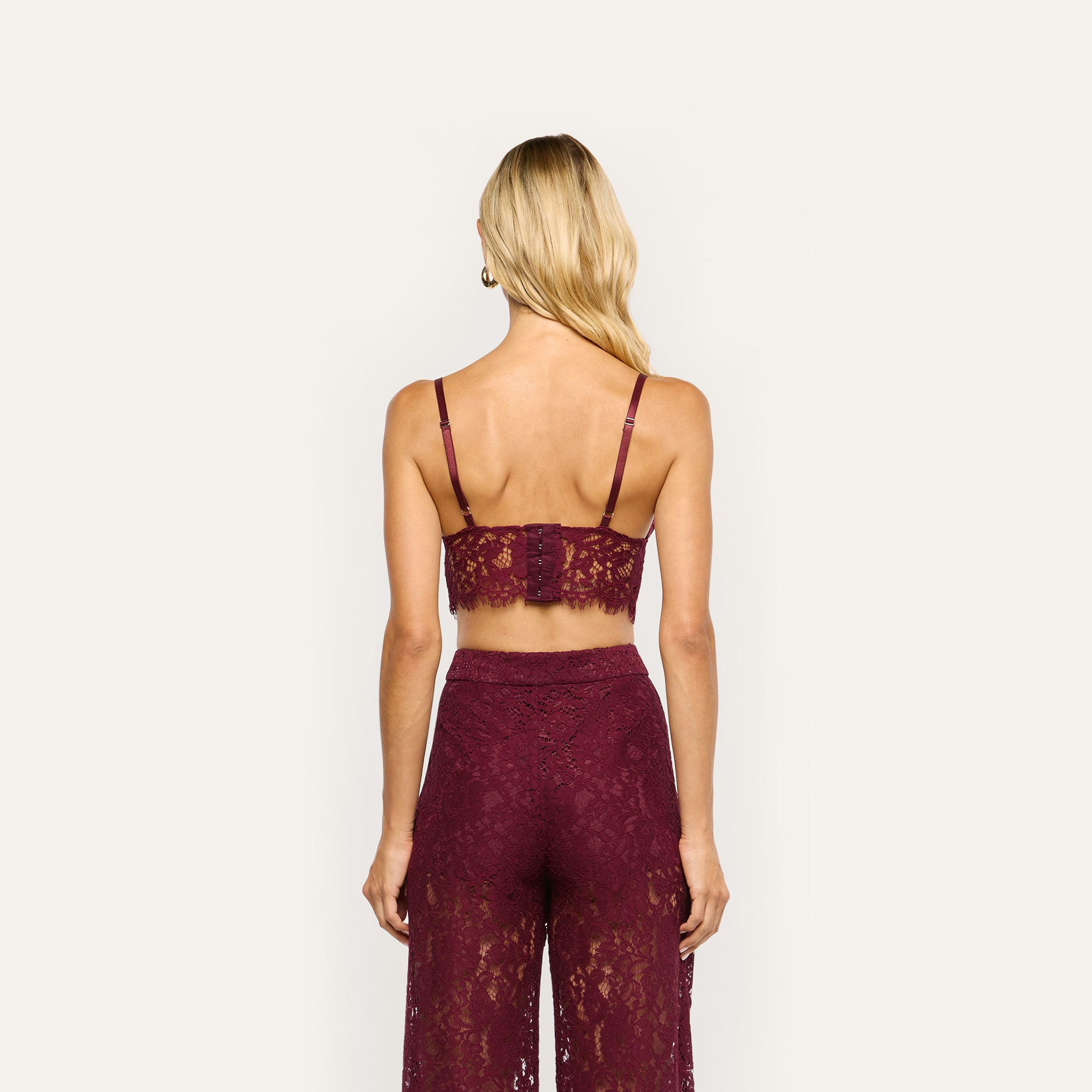 A person seen from behind wearing an Aislin Lace Bralette and matching trousers in burgundy sheer stretch lace with scalloped edges.