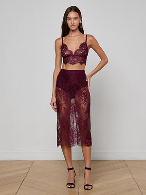 A woman with long hair and a neutral expression wears the maroon Aislin Lace Bralette by L'AGENCE and a matching sheer high-waisted lace skirt, paired with black high-heeled sandals, while standing against a plain wall.