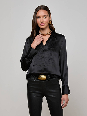 A woman with long brown hair wears the L'AGENCE Divya Silk Shirt, paired with black leather pants, gold hoop earrings, layered necklaces, and a gold belt buckle, standing against a plain light gray background.