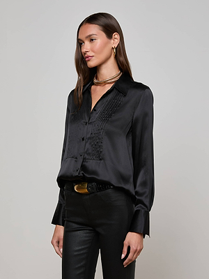 A woman poses against a plain backdrop, wearing the L'AGENCE Divya Silk Shirt in black with pleated details and satin buttons, paired with black pants, gold hoop earrings, layered gold necklaces, and a black belt with a gold buckle.