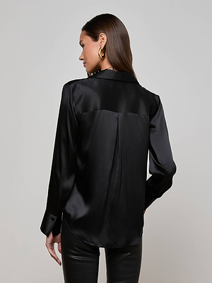 A woman with long brown hair wears gold hoop earrings, black leather pants, and the Divya Silk Shirt by L'AGENCE as she stands facing away from the camera against a plain light gray background.