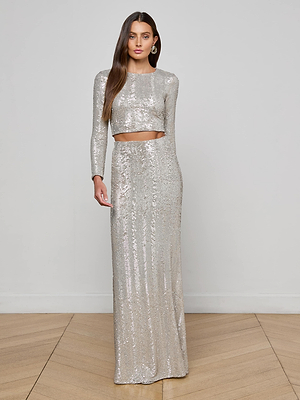 A woman with long brown hair and drop earrings stands on a wooden floor, wearing the L'AGENCE Rogan Sequin Cropped Top with long sleeves and a matching high-waisted skirt. A chic, silver party look against a plain background.