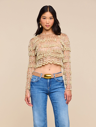 A woman stands against a beige background, looking at the camera with a neutral expression. She wears the Rogan Pearl Fringe Top—a sheer, long-sleeve crop top adorned with delicate pearl fringe—paired with blue jeans. Her chic ensemble captures modern glamour and echoes NYFW Fall 2026 trends.