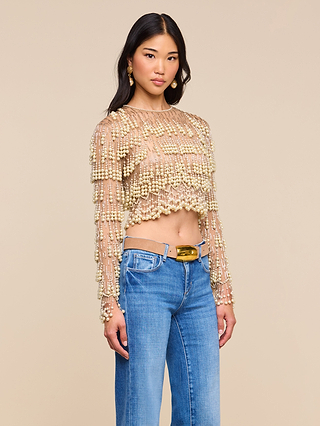 Wearing the Rogan Pearl Fringe Top in sheer beige, adorned with gold beads, paired with high-waisted blue jeans, she poses against a plain beige background—a look perfect for NYFW Fall 2026.