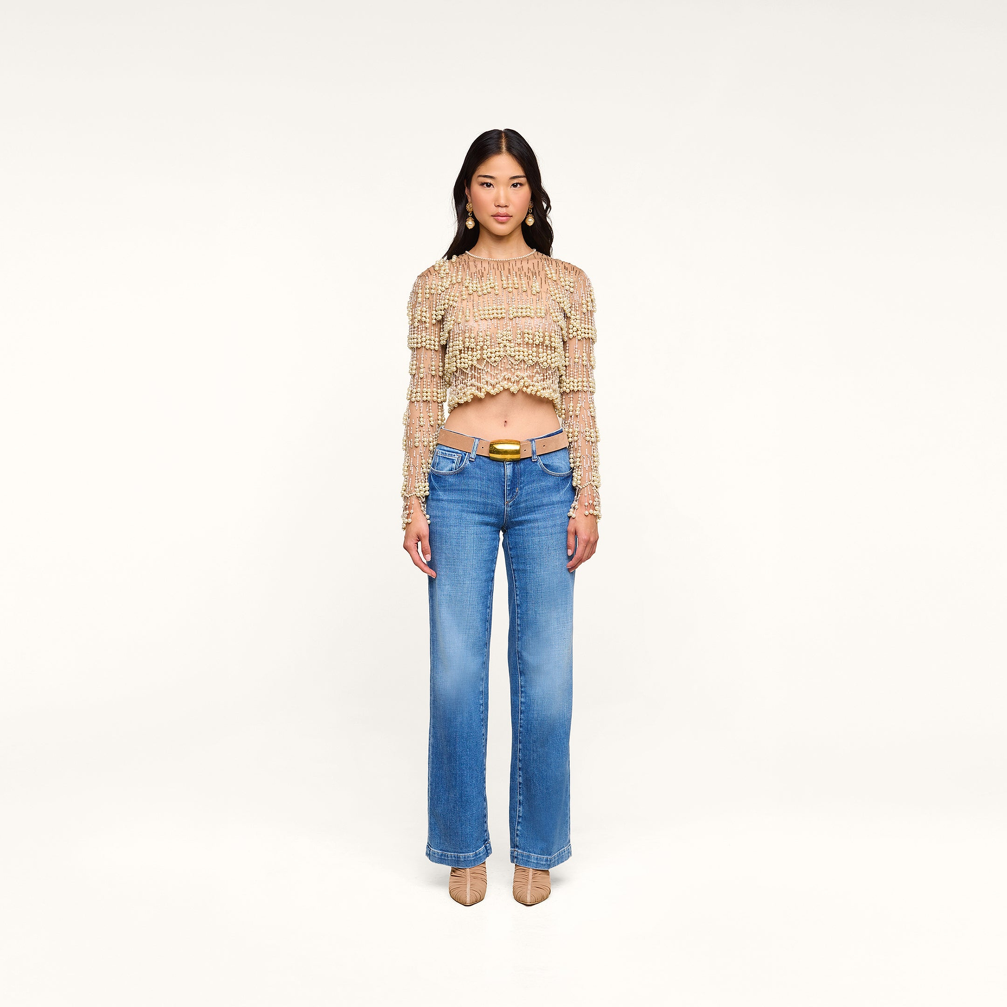 Woman stands facing forward wearing the Rogan Pearl Fringe Top, a mesh long sleeve beige crop top, paired with blue wide-leg jeans with a belt and tan heels, against a plain white background.