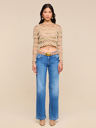 Woman standing against a beige background, wearing the Rogan Pearl Fringe Top—a textured crop top with pearl fringe and long sleeves—paired with blue high-waisted jeans, a tan belt, and beige pointed-toe shoes. With her long dark hair and confident forward-facing pose, she channels NYFW Fall 2026 vibes in this effortlessly chic ensemble.