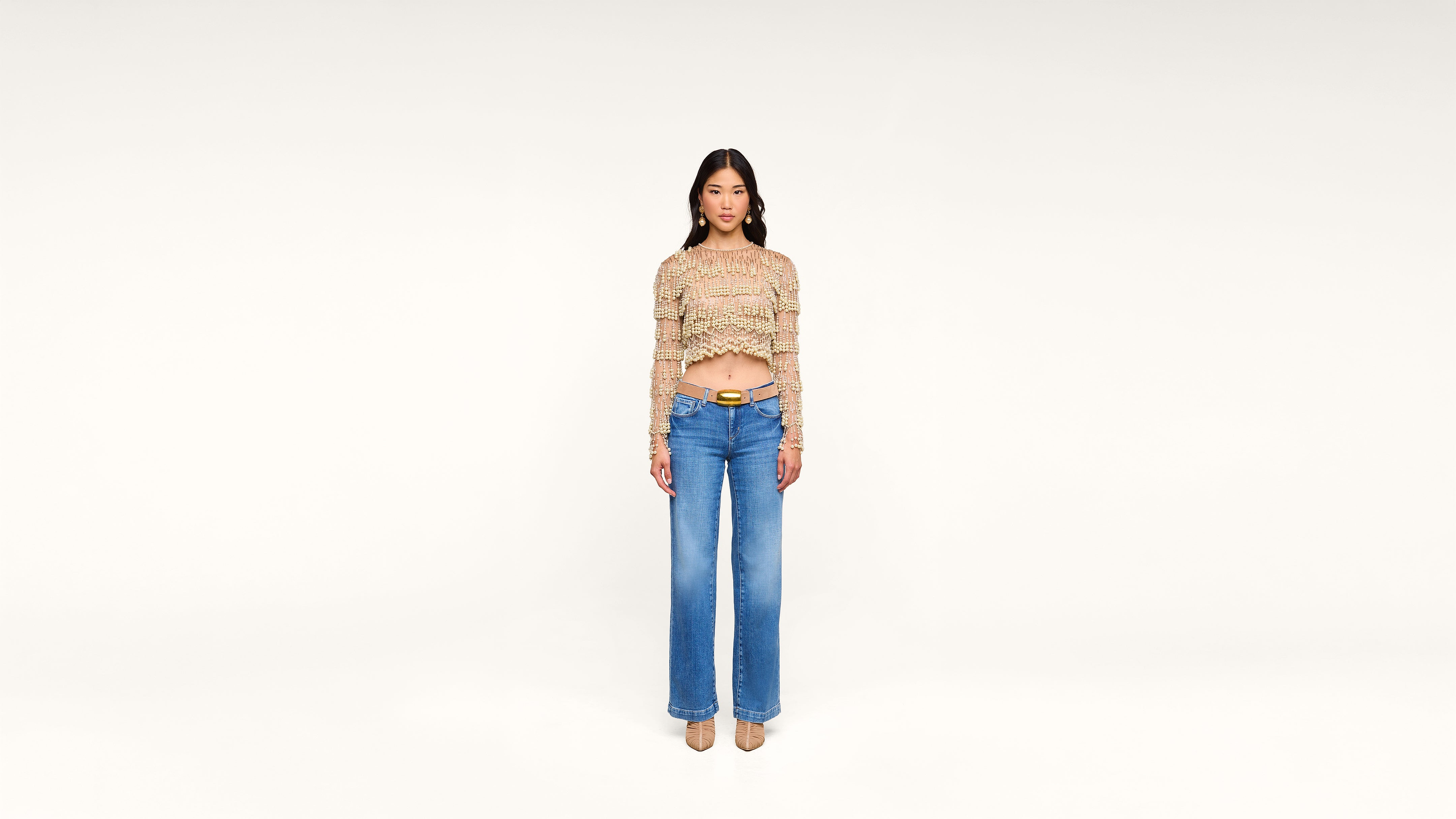 A woman stands facing forward against a plain white background, wearing a long-sleeve, multicolored crop top, high-waisted blue jeans, a tan belt, and beige shoes.