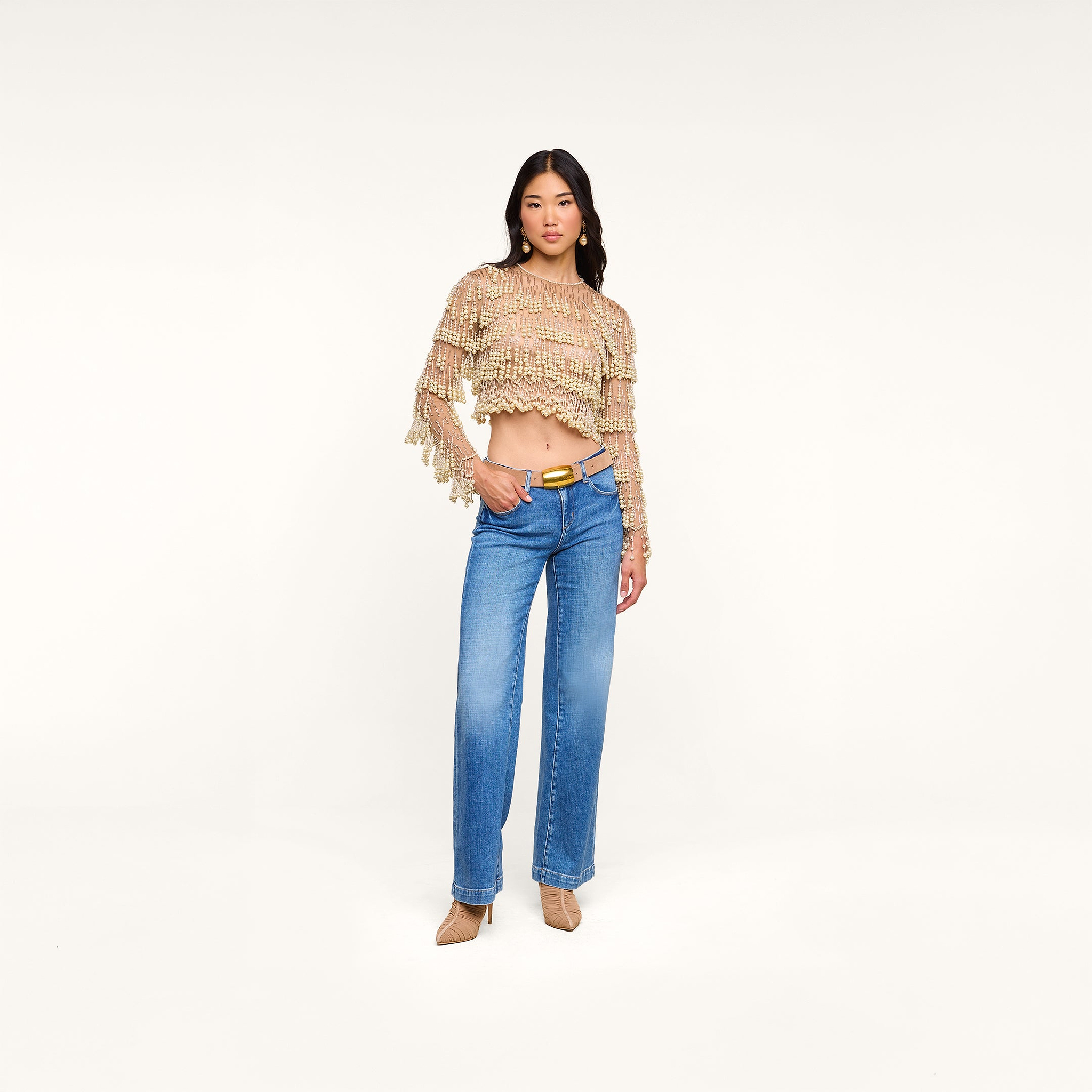 A woman poses in the Rogan Pearl Fringe Top, a beige mesh long sleeve crop top, paired with blue high-waisted jeans, a tan belt, and tan heels against a plain white background.