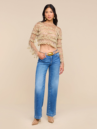 A woman stands against a beige background wearing the Rogan Pearl Fringe Top with loose sleeves, blue high-waisted jeans, a beige belt, and pointed-toe beige heels—a chic look reminiscent of NYFW Fall 2026.