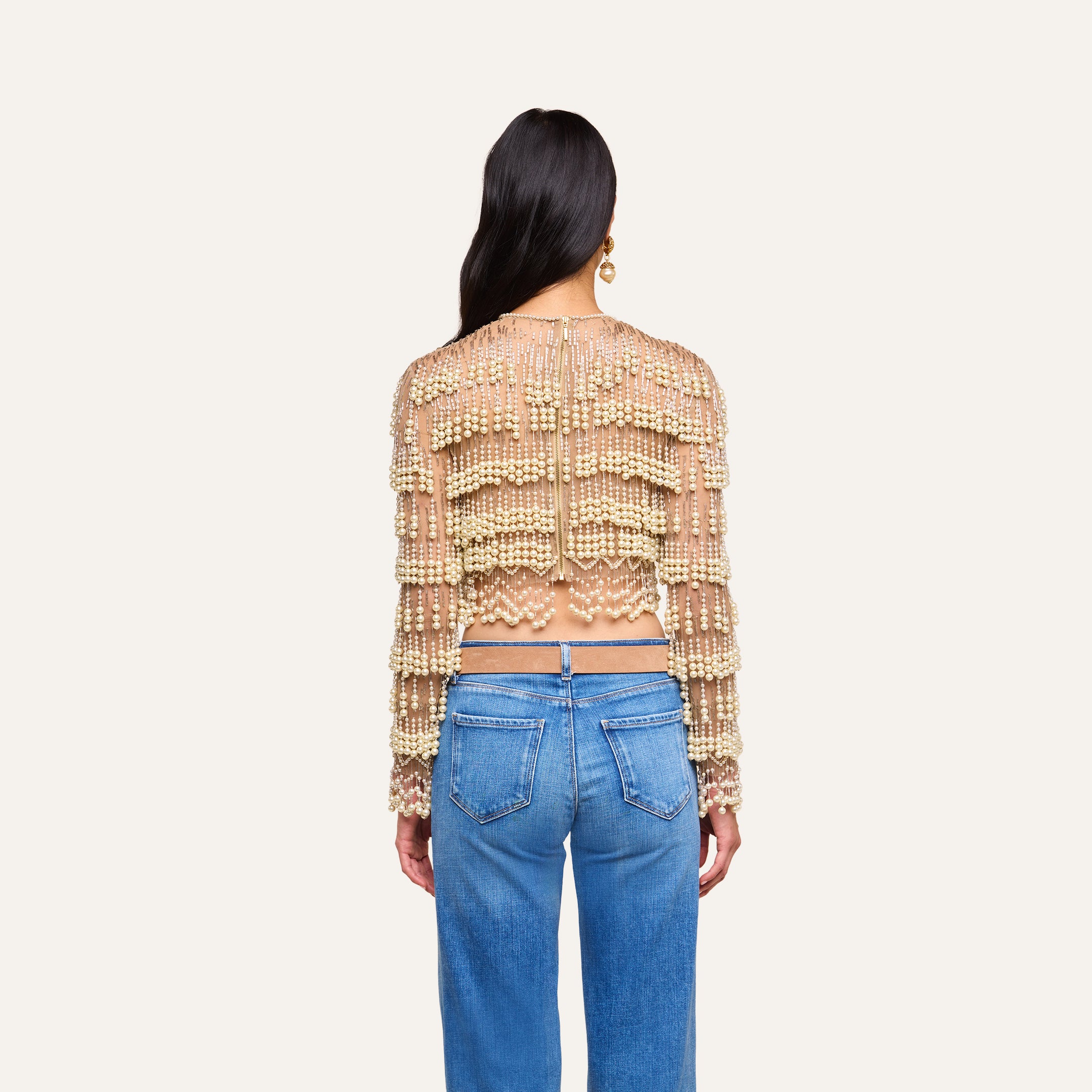 A person with long dark hair, dressed in the Rogan Pearl Fringe Top and blue jeans, stands with their back to the camera against a plain, light background.
