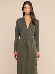 A woman stands facing forward, wearing the Isla Shirt in a long-sleeve olive green featherweight twill, featuring button details, a drawstring waist, and large front pockets. She has long brown hair and a neutral expression. The background is plain off-white.