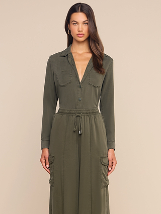 A woman wears the Isla Shirt, an olive green featherweight twill long-sleeve button-up dress featuring front pockets and a drawstring waist, standing against a plain beige background.