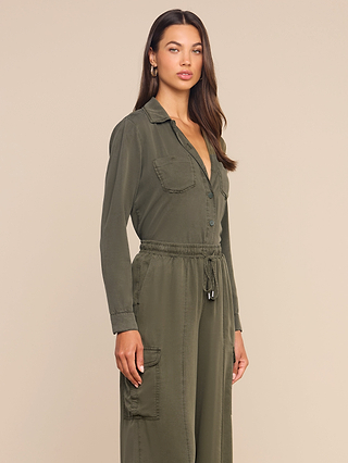 A woman wears the Isla Shirt, an olive green long-sleeve utility top crafted from featherweight twill with chest and cargo pockets, standing against a beige background and looking confidently at the camera.