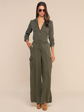 A woman wearing the Isla Shirt in olive-green featherweight twill with long sleeves, a button front, drawstring waist, and cargo pockets, paired with brown boots and sunglasses, stands against a plain beige background.